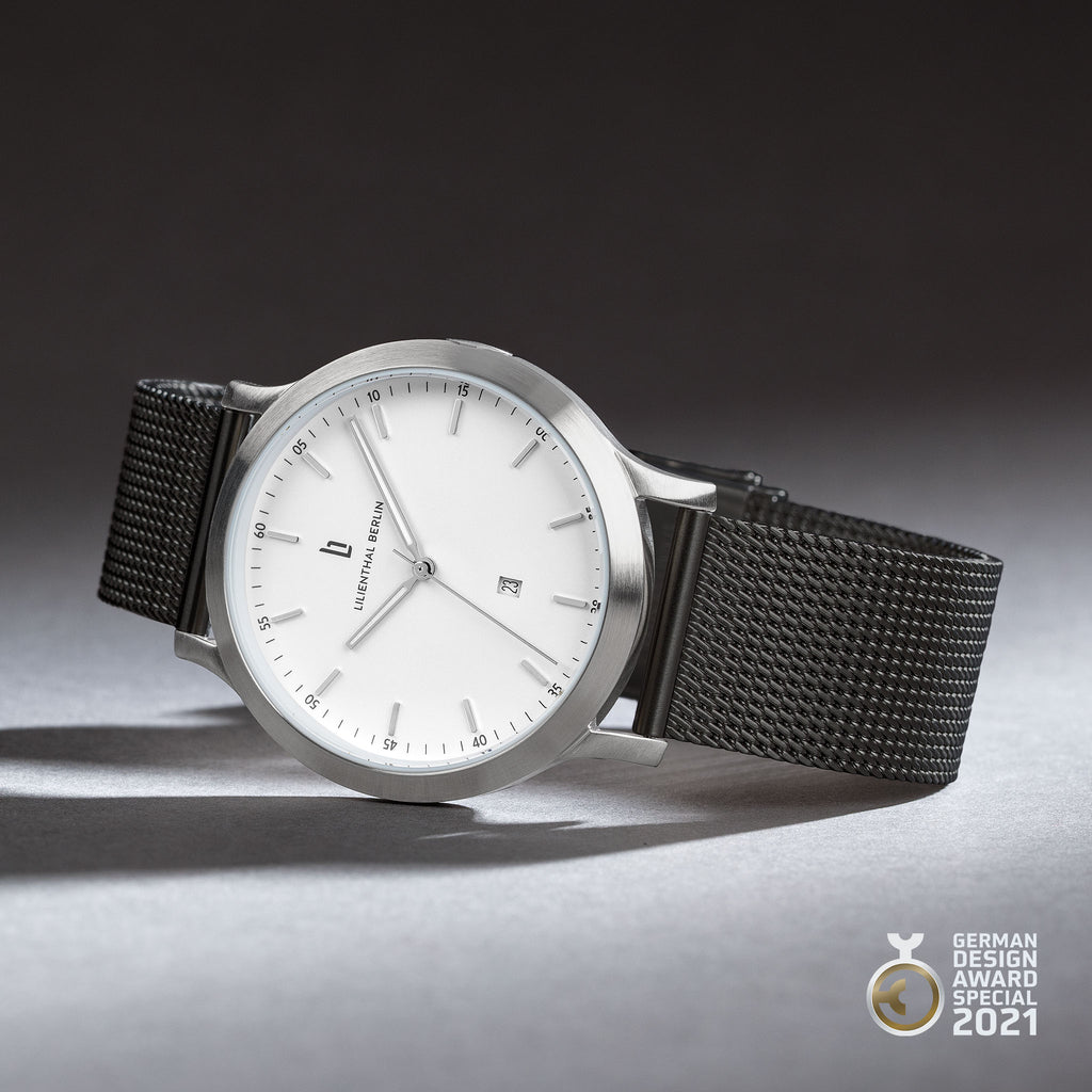 Minimalist silver and white wristwatch with black mesh strap, photographed on a gray surface