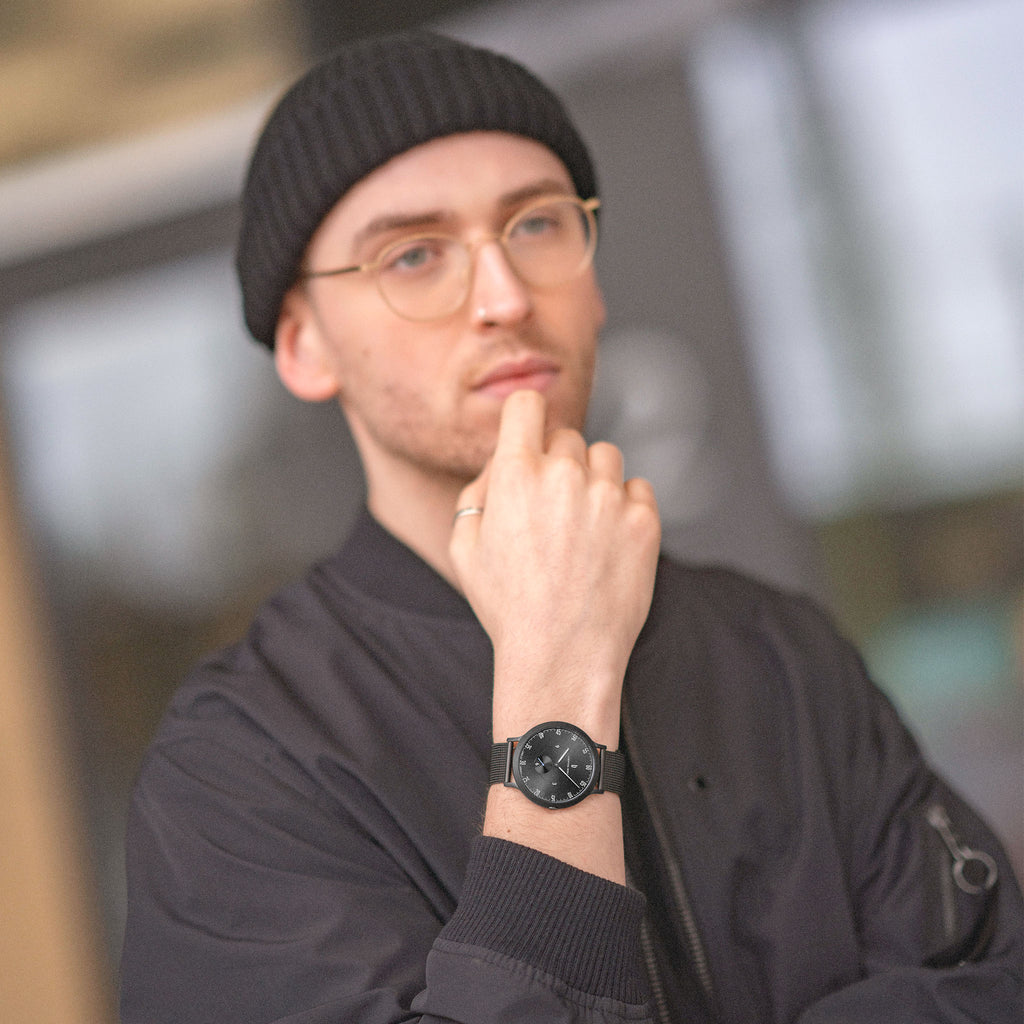 Man wearing a black minimalist wristwatch, black jacket, glasses, and beanie outdoors.