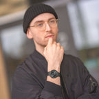 Man wearing a black minimalist wristwatch, black jacket, glasses, and beanie outdoors.